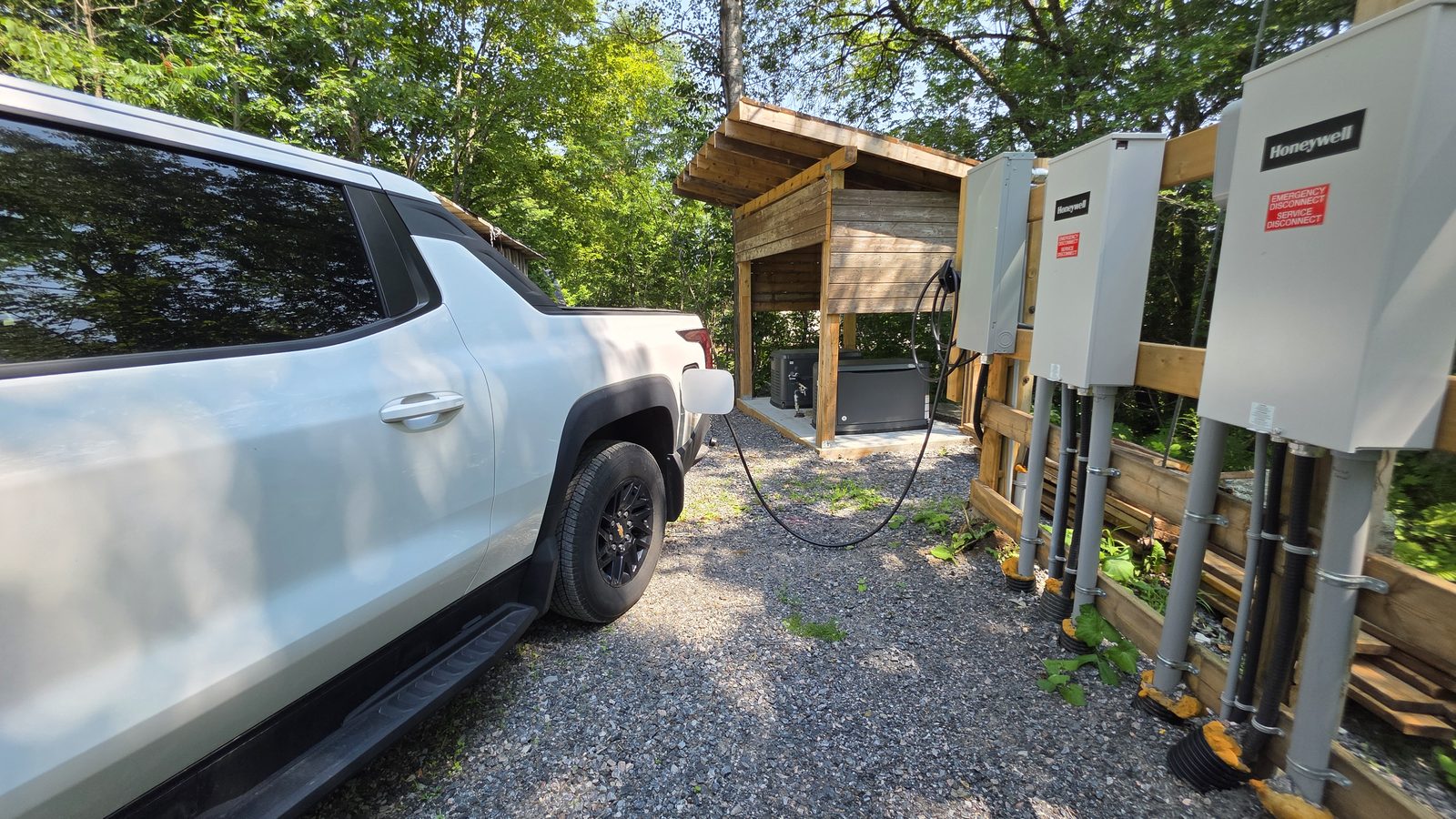 A white Chevrolet Silverado EV plugged in and charging on a gravel driveway. Behind it a timber shed holding two propane standby generators, and on the right their transfer switches mounted on posts.